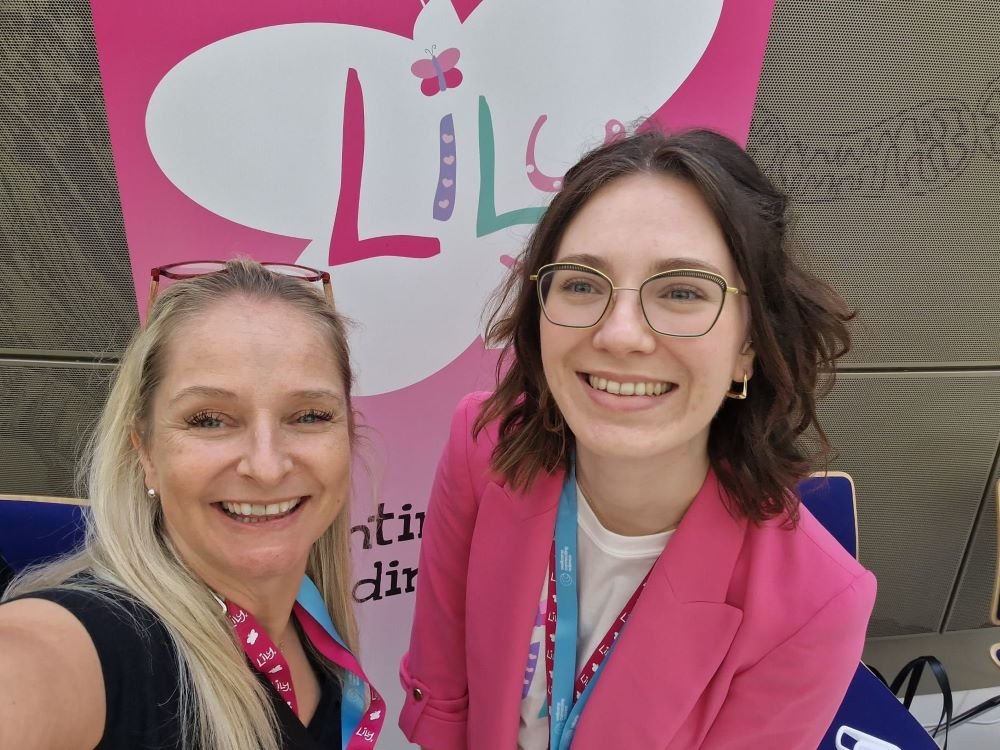 Two ladies smiling and standing in front of a pink Lily Foundation banner