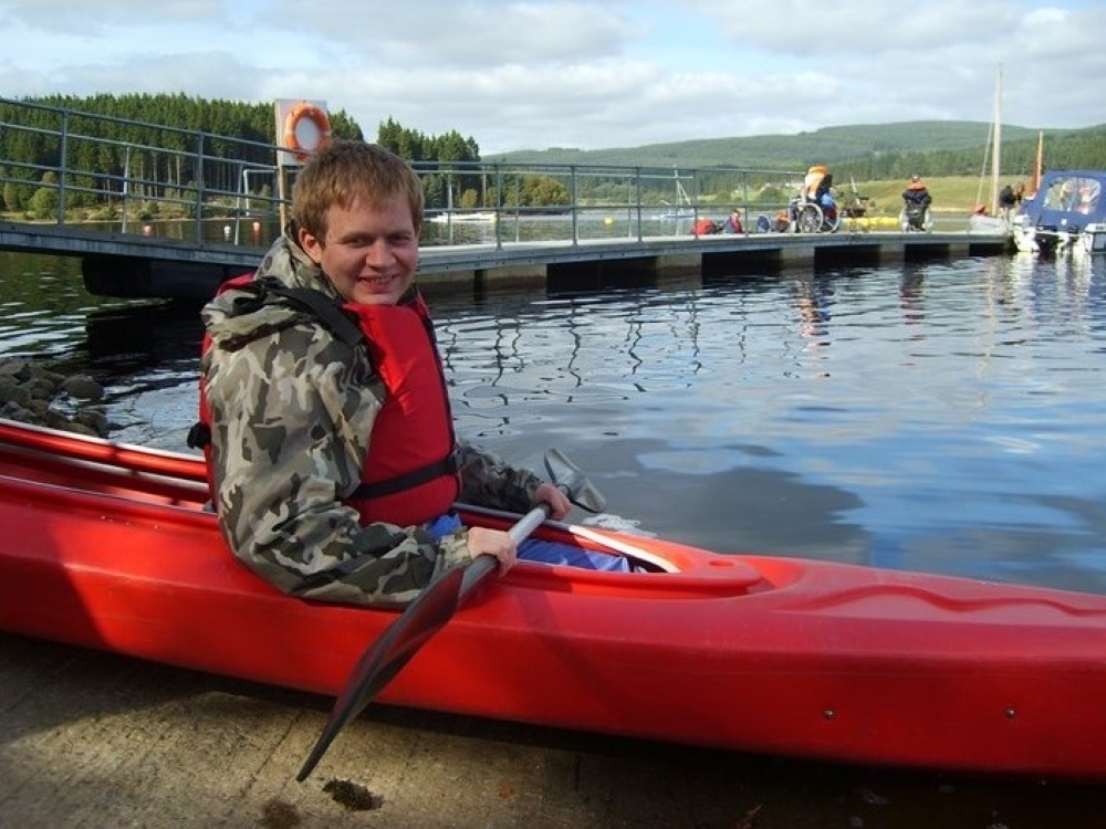A smiling man in a red kayak on a lake holding the paddle