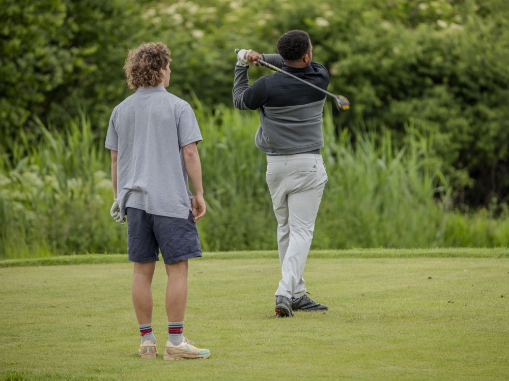 A man swings his golf club on the course as another man stands nearby watching