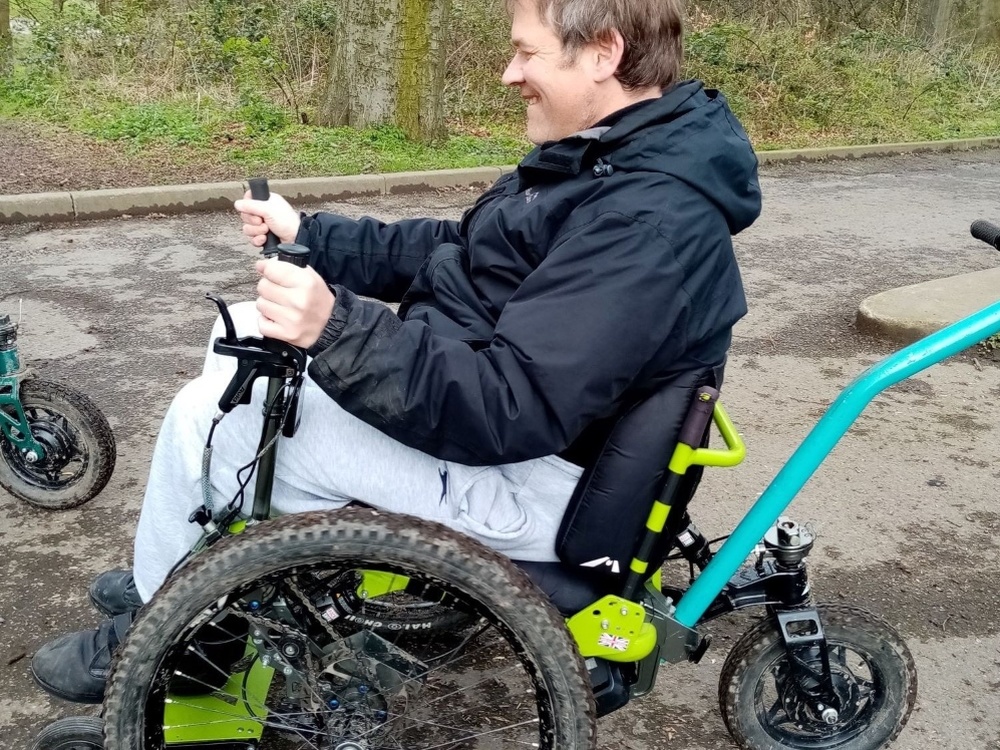 A side on view of a smiling man on a muddy Mountain trike using its control levers