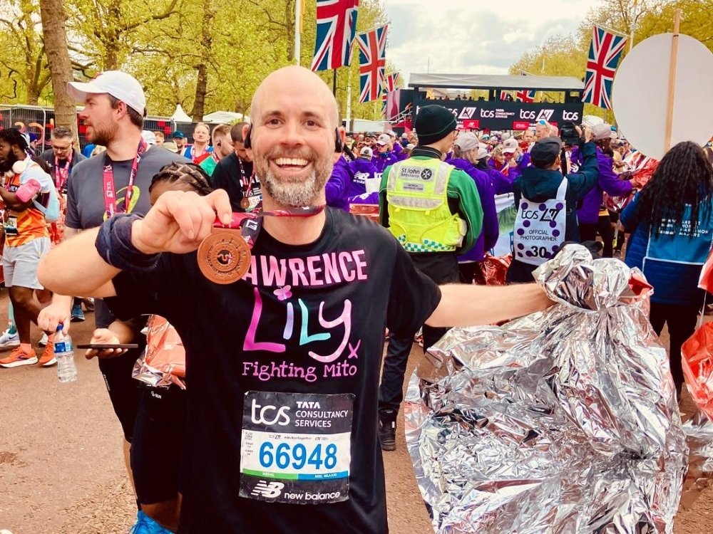 A man in a Lily vest smiling and holding a London Marathon finishers medal up