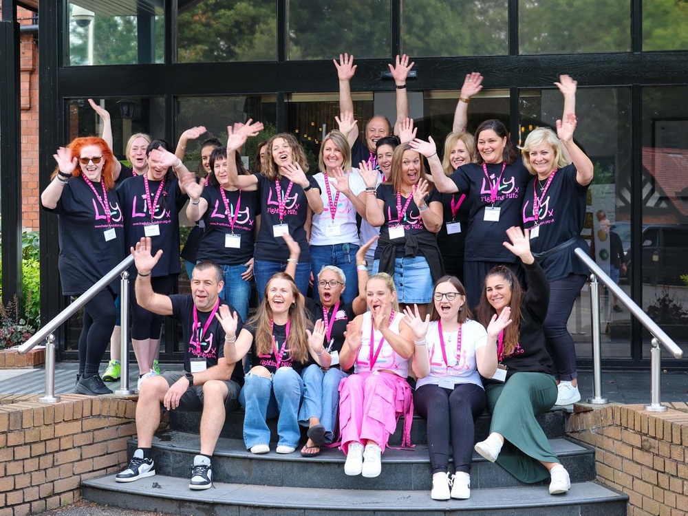 A large group of adults, all wearing Lily t-shirts, are grouped around the front steps of a hotel, smiling and waving