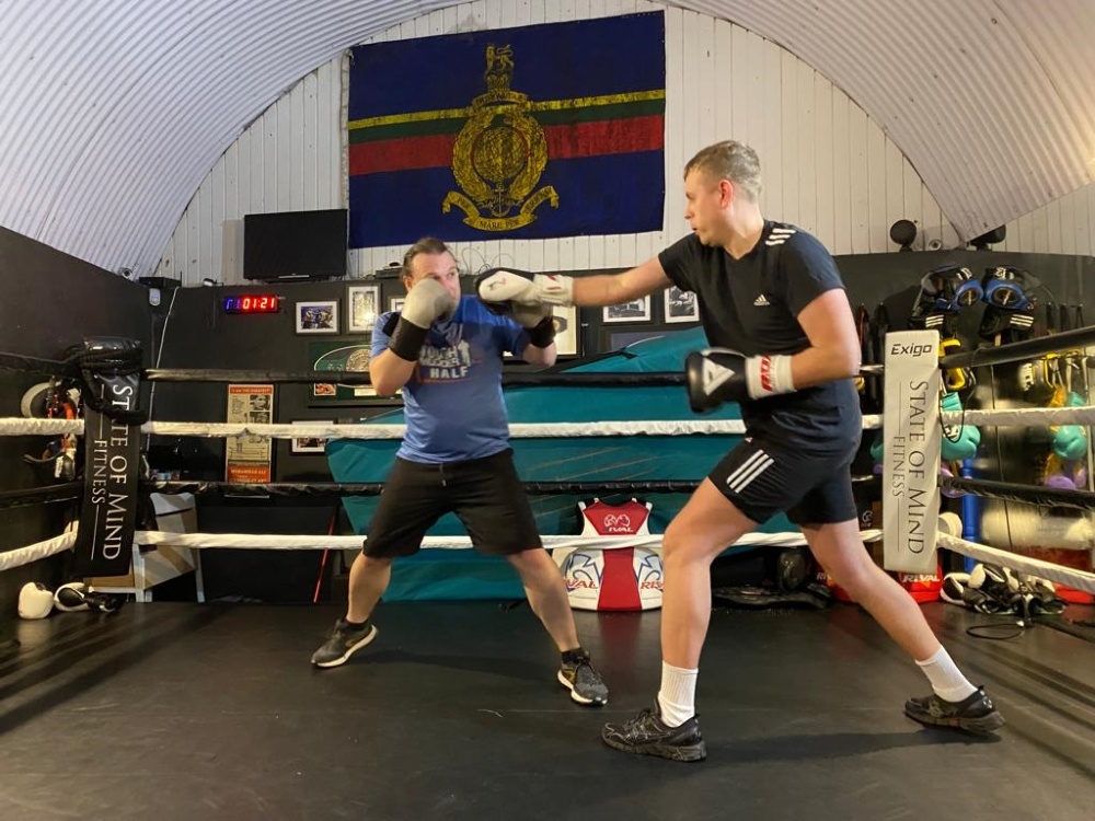 Two men in a boxing ring wearing boxing gloves. One has his hands up guarding his face the other is swinging a punch