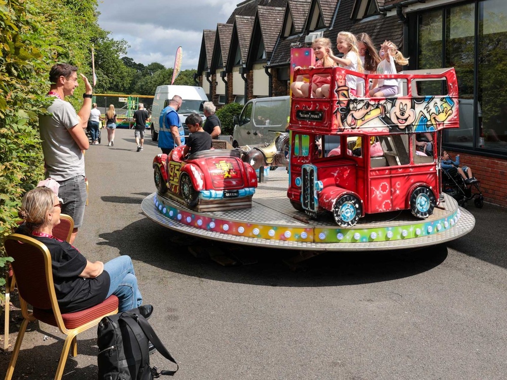 A group of young girls ride a carousel, waving, while a man and lady watch and wave back at them from the side