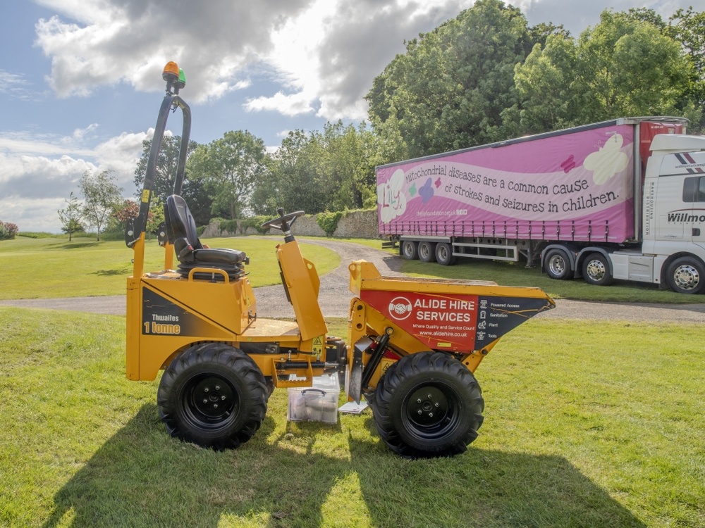 A dumpster on a golf course in front of a large lorry with a Lily Foundation logo