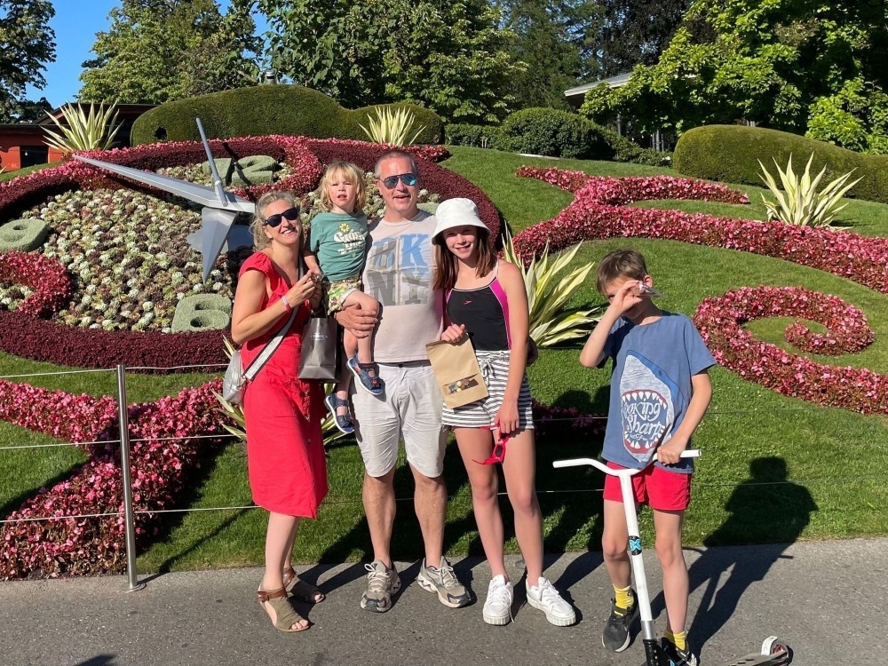 A family stand together in the sun in front of gardens with a clock behind