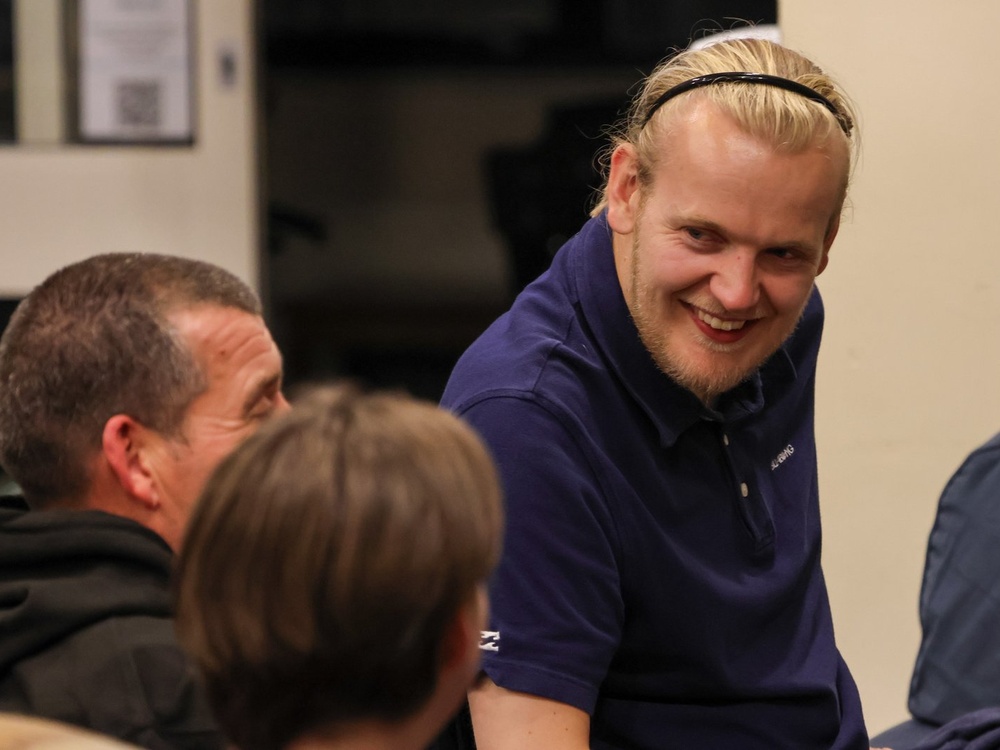 A man with blonde hair pushed back by a hairband is turning laughing at people to his left