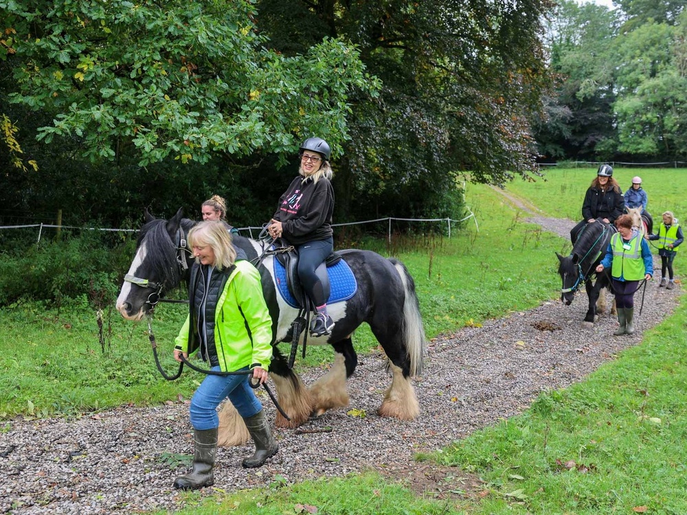 A lady sitting on a horse is being led along a path by another lady with two more horses behind