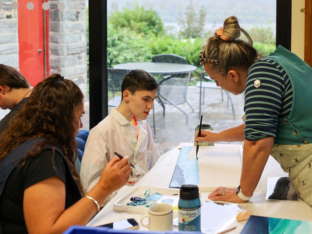 A young man bends over a painting while a lady leans in with a paintbrush and another lady looks on