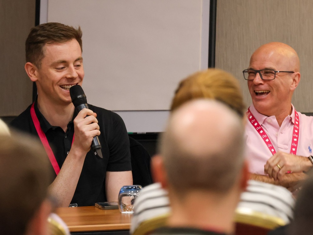 Two men are seated behind a desk, one speaking into a microphone, to a group of adults in front of them