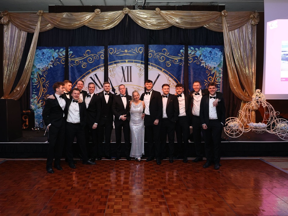 A group of men in black tuxedos and a lady in a silver dress standing in a row in front of a stage with a large clock