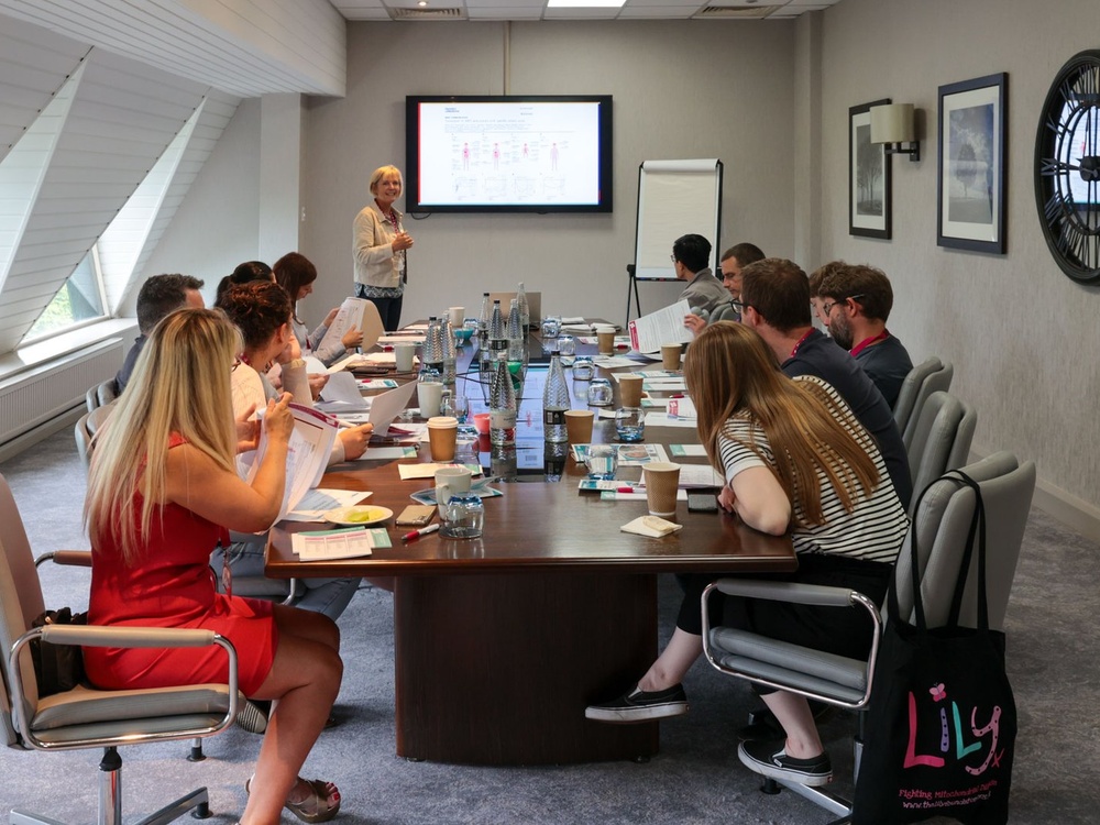 A group of people sit around a long oblong table looking at someone presenting at a screen in front of them