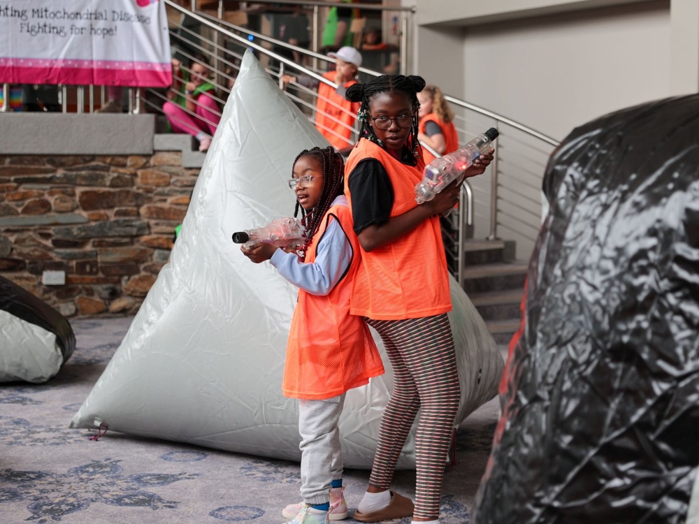 Two children holding laser guns and wearing orange bibs are standing back to back, poised to shoot