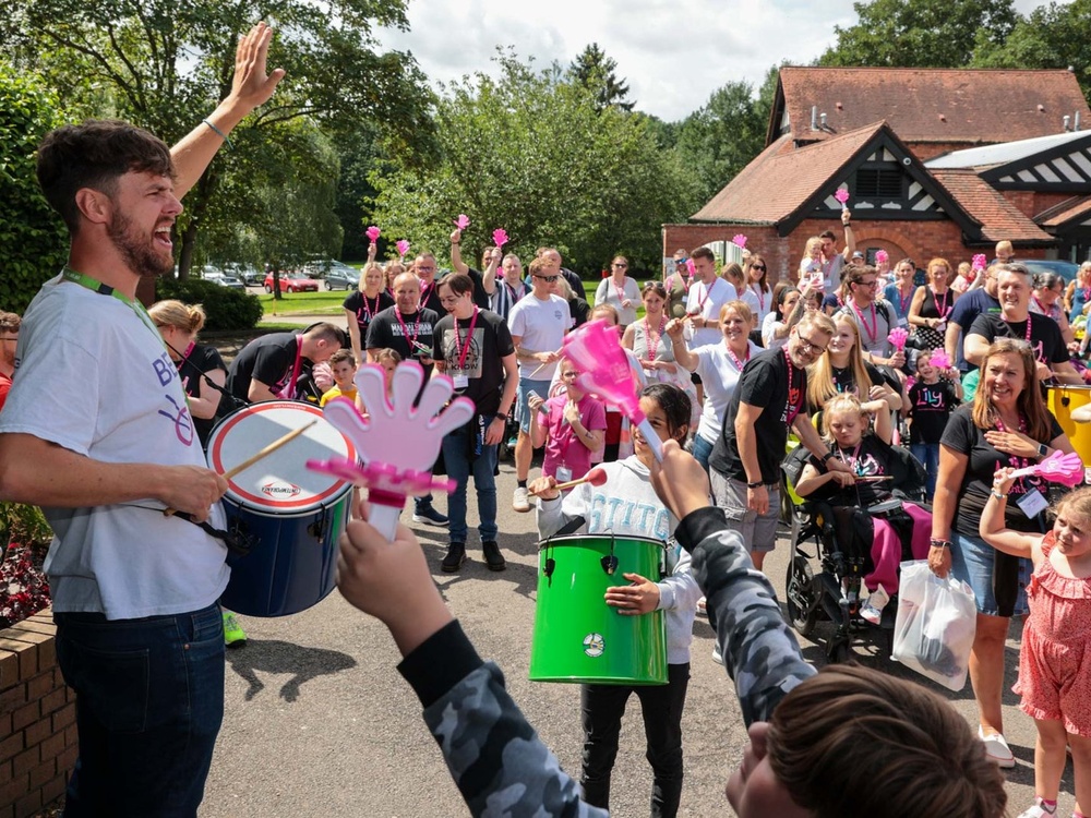 A large number of people, many in Lily t-shirts, are standing outside a hotel clapping while a man bangs a drum at the front