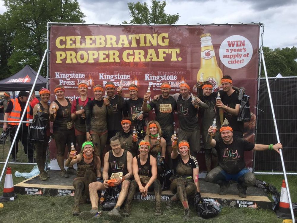 Tough Mudder 2024 - Locations to choose from across the UK - A group of people wearing Lily Foundation tops, covered in mud and standing on a wooden platform