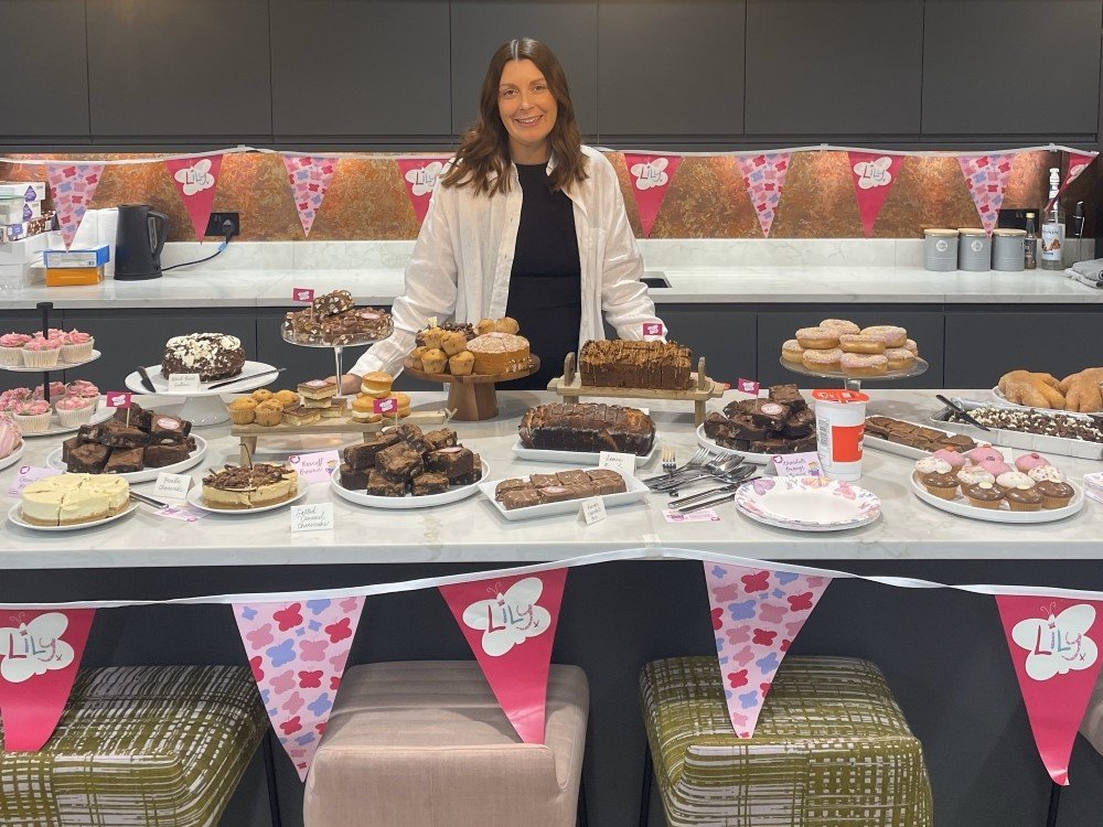 A lady standing behind a table laid with lots of cakes and Lily Foundation banners and leaflets