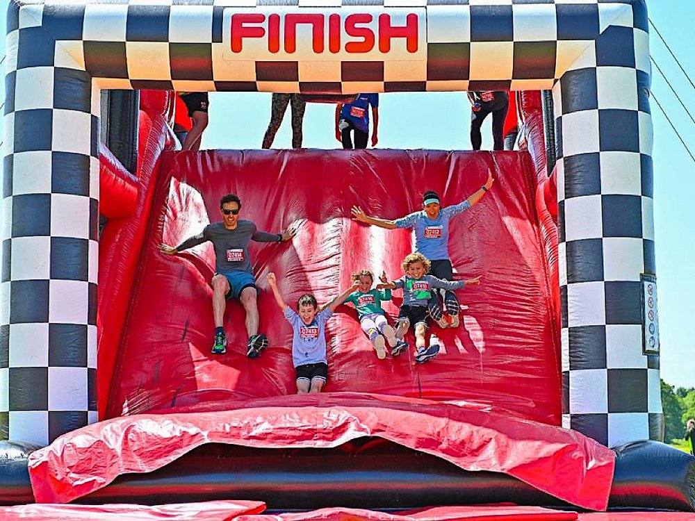 The Inflatable 5k Run Series - Adults and children with arms raised sliding down a large inflatable finish line slide