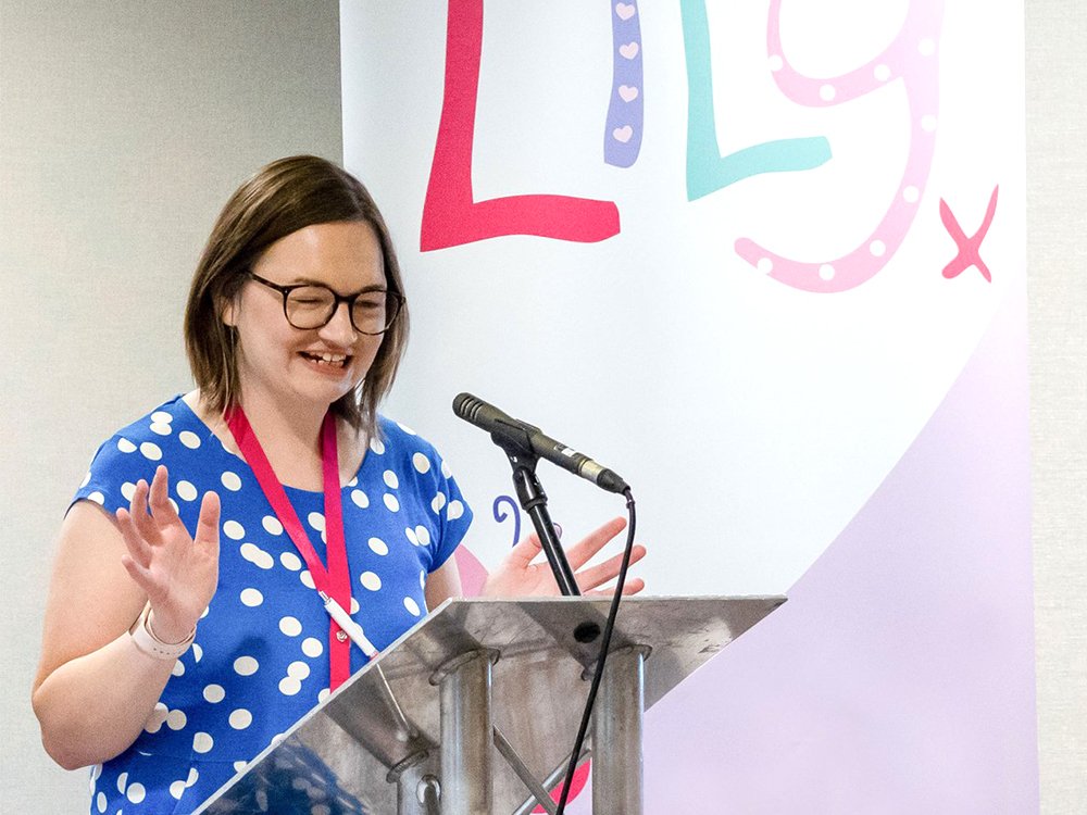 Lady standing at a lectern speaking into a microphone with a Lily banner in the background