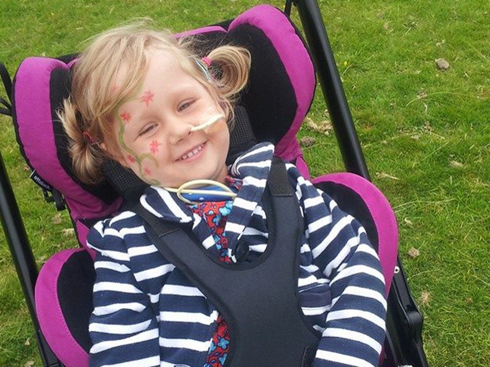 Young girl smiling wearng a blue and white stripy top, sitting in her wheehchair with a feeding tube in her nose