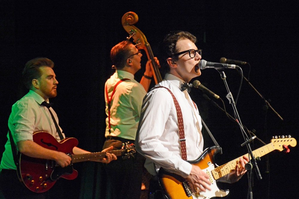 Man in a white shirt and braces, playing a guitar in front of a microphone. Other members of the band are in the background