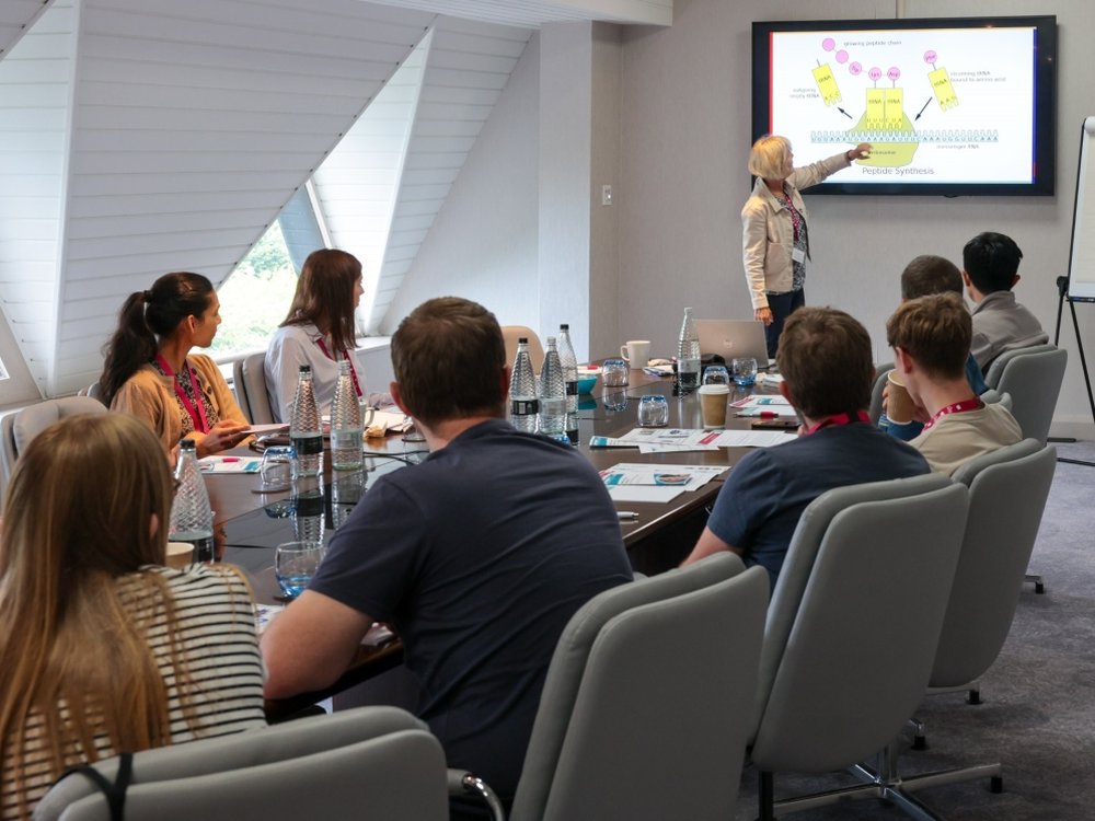 A woman pointing at a large screen, standing in front of a group of people sitting around a table