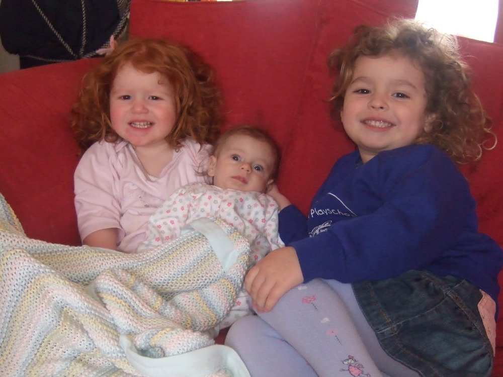 Baby Lily on a red sofa with her big sisters Katie and Rosie