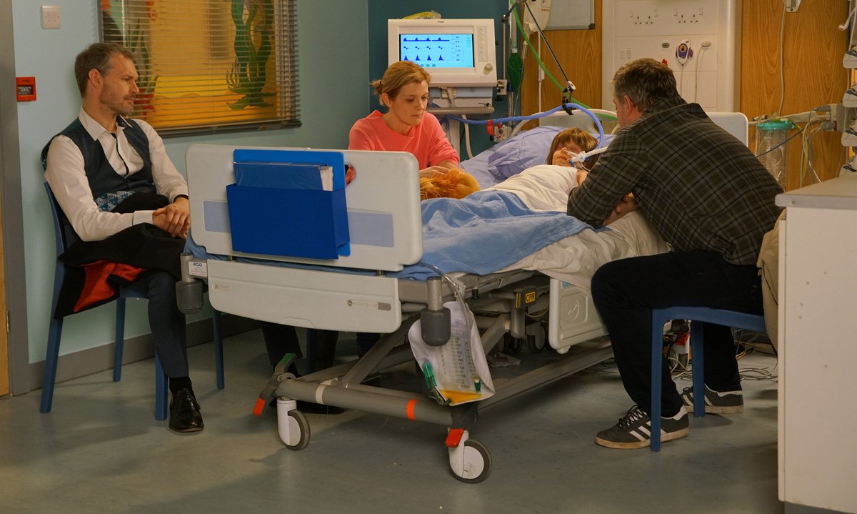 two men and a woman sit around a hospital bed. In it is a little boy asleep connected to a breathing tube