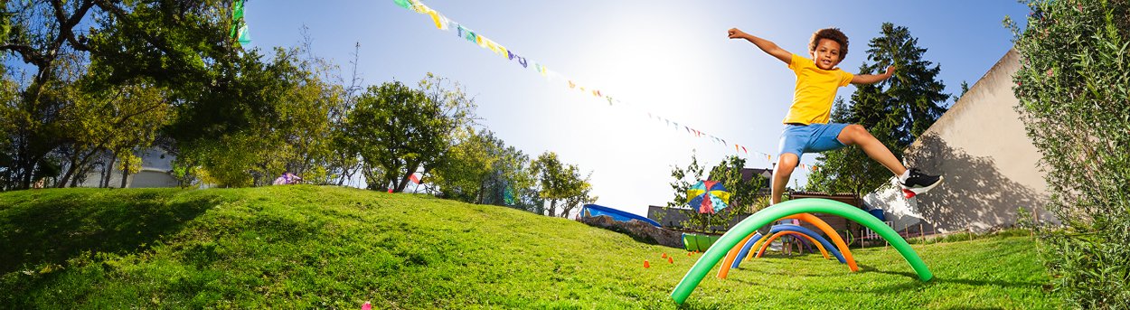 A sunny garden with colourful pool noodles stuck into the ground to make arches. A young boy is leaping over the hurdles