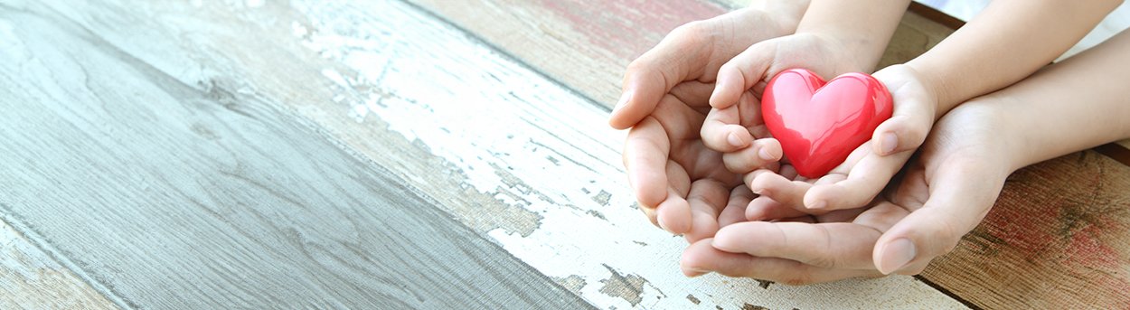 Child's cupped hands holding a red heart, nestled within cupped adult hands