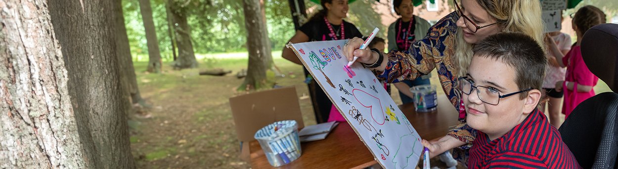 Young boy with mito in the woods with a helper, drawing on a white canvas, symbols of love with big colourful marker pens