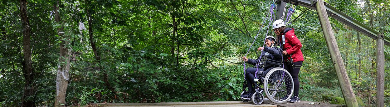 Young adult in her wheelchair being pushed by her mum over a forest assault course