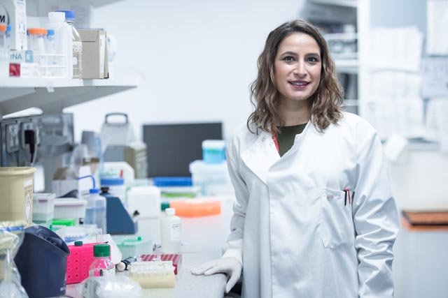 A woman stand next to a laboratory bench covers in various bottle and containers. She is wearing a white lab coat and has shoulder length brown wavey hair