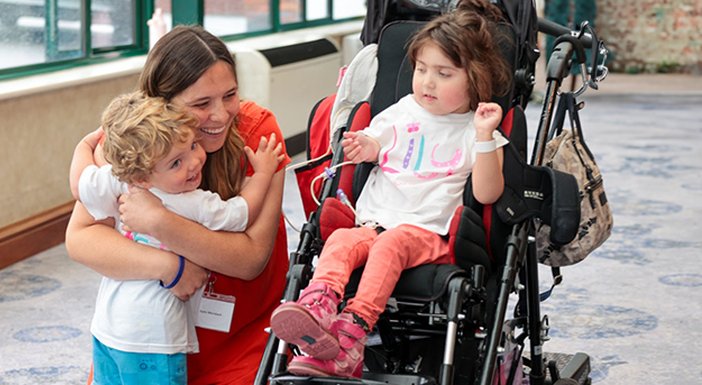 Mum crouched down smiling and  cuddling her child in a lily Tshirt, whilst another child in a pushchair is smiling