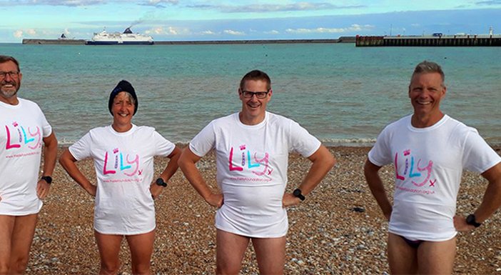 5 Lily fundraisers standing on a pebble beach in  swimming shorts and Lily T-shirts, ready to take the plunge!