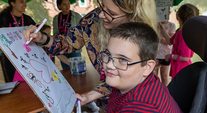 Young boy with mito in the woods with a helper, drawing on a white canvas, symbols of love with big colourful marker pens