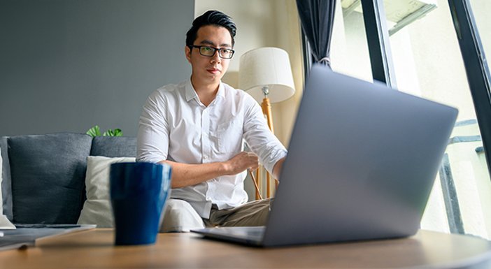 Man sitting on the end of a grey sofa, engaging with someone via his open laptop