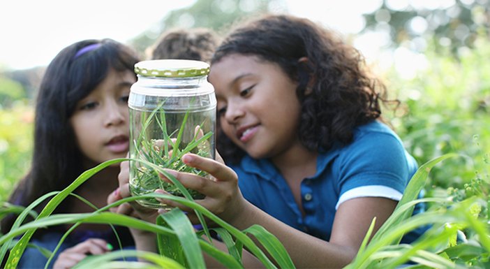 2 girls lying in long grass, looking at a jam jar with leaves and bugs inside