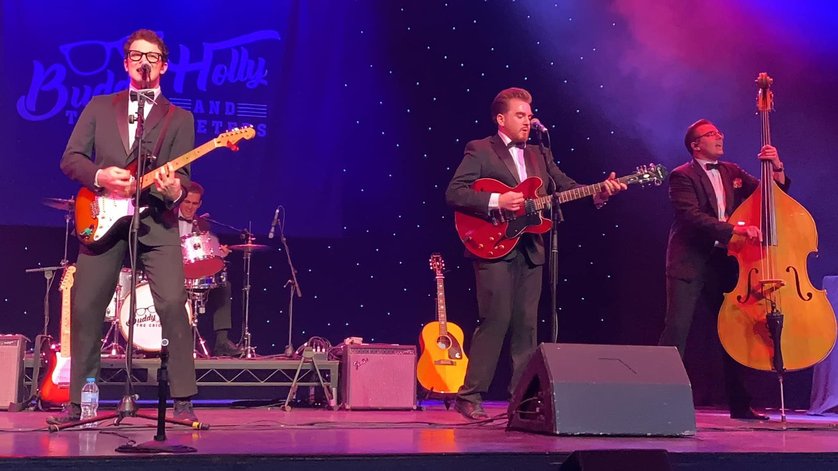 three men in suits and bow ties stand on stage. Two are playing guitar and singing into micrphones. ONe is playing a double base. Behind then is a mann sat behind a set of drums