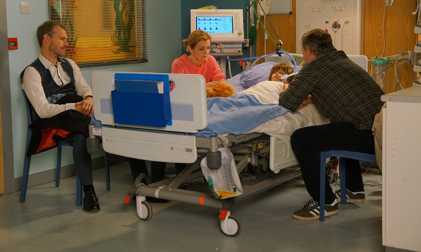 two men and a woman sit around a hospital bed that has a little boy in it. He is asleep and connected to a breathing tube