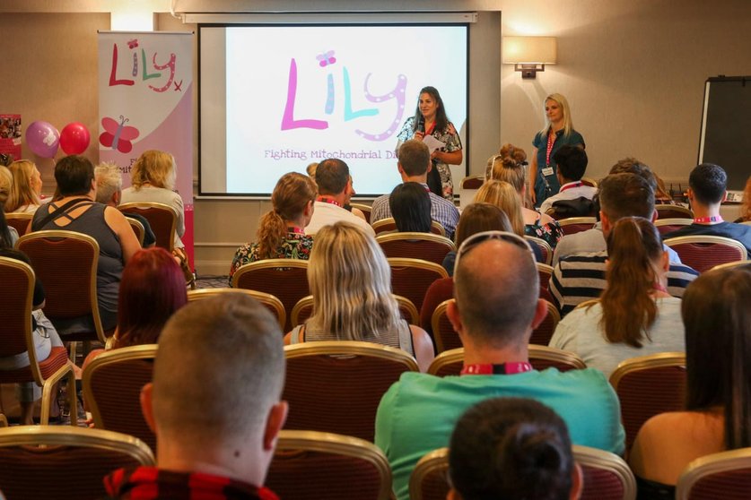 A room full of people sat on rows of chair faceing a screen with The Lily Foundation on it. Looking out towards them are two women