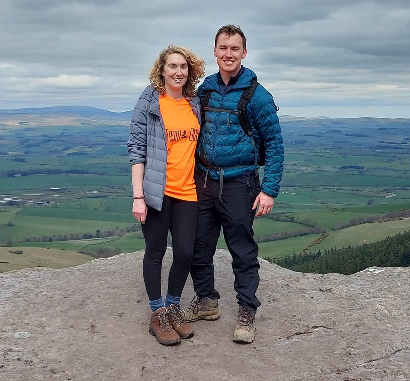 A woman and a man stood together with one arm round each other on a hill with green fields behind them.