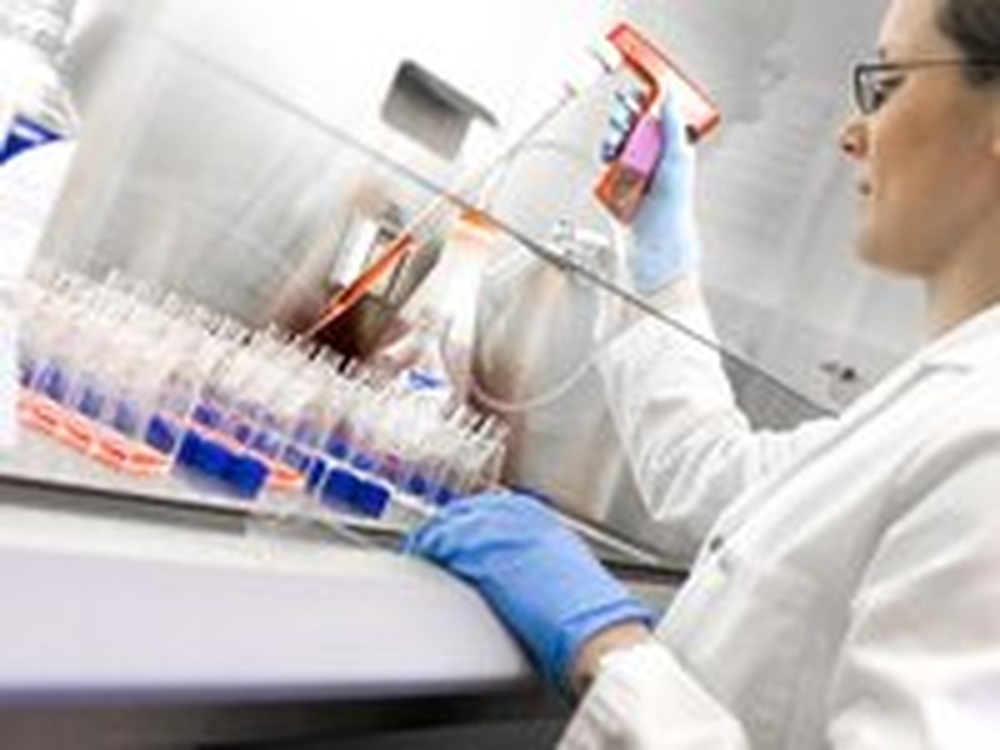 A women in safty googles and lab coat uses a micro pipette at a bench covered in viles