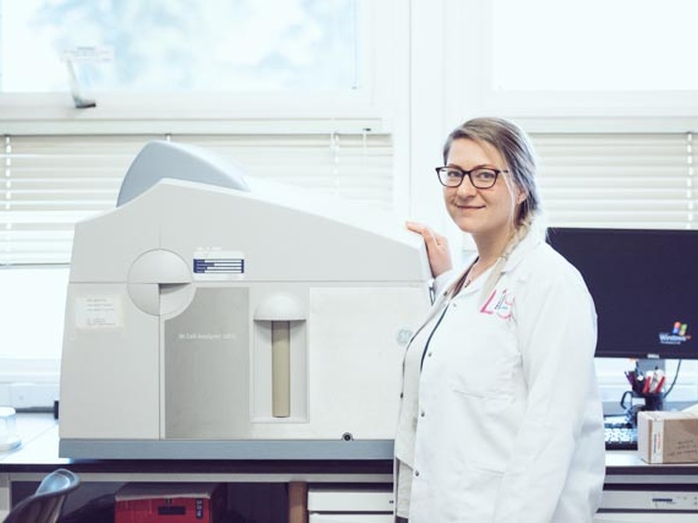 A women in a white lab coat with her brown hair tied back with glases stands next to a piece of scientific equipment