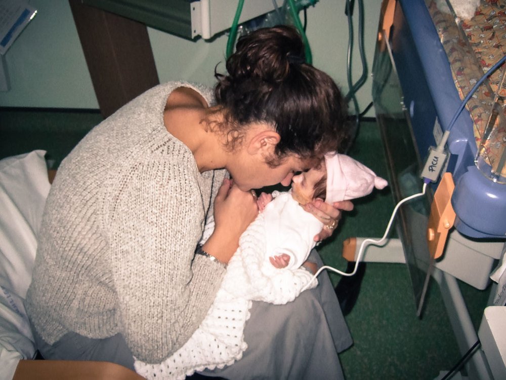 A Women sat in a chair holding a small baby. They are touching noses. The baby is attached by wires to a medical device