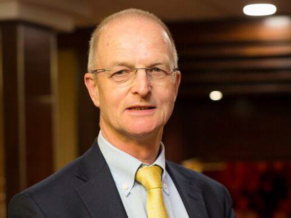 A head shot of a man with glasses in a dark suit and yellow tie