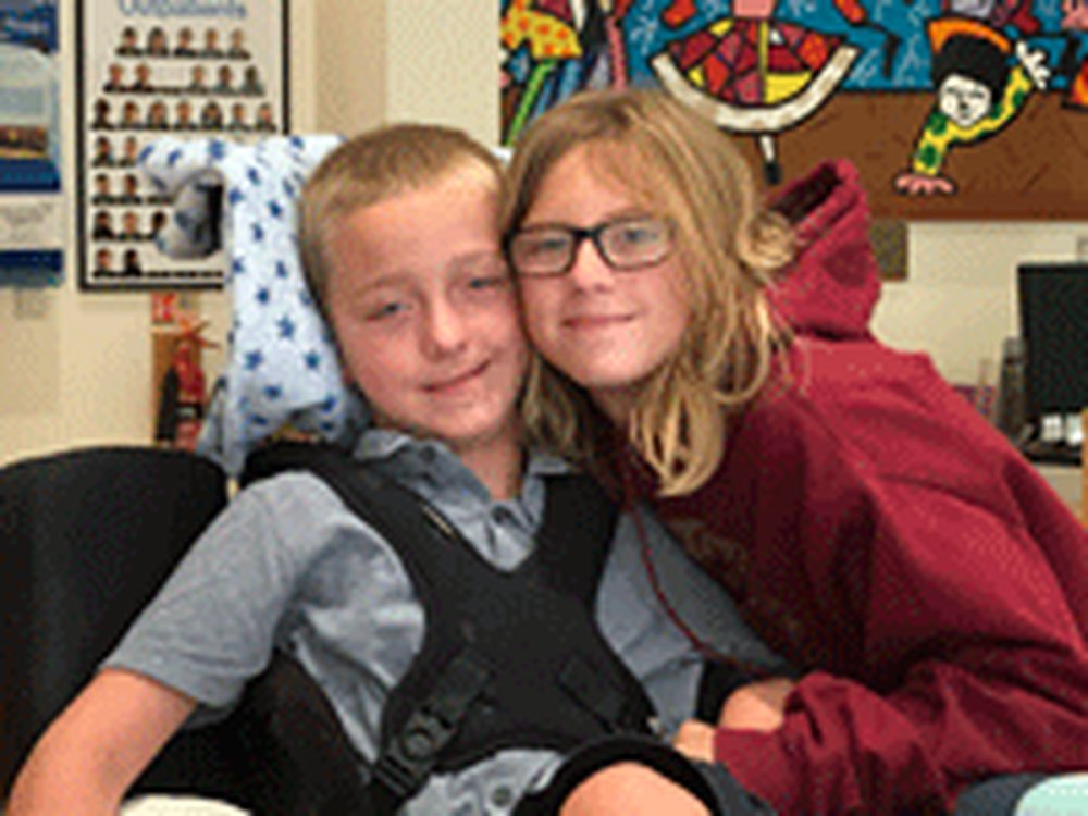 A boy in a wheelchair sits cheek to cheek with a little girl with glasses