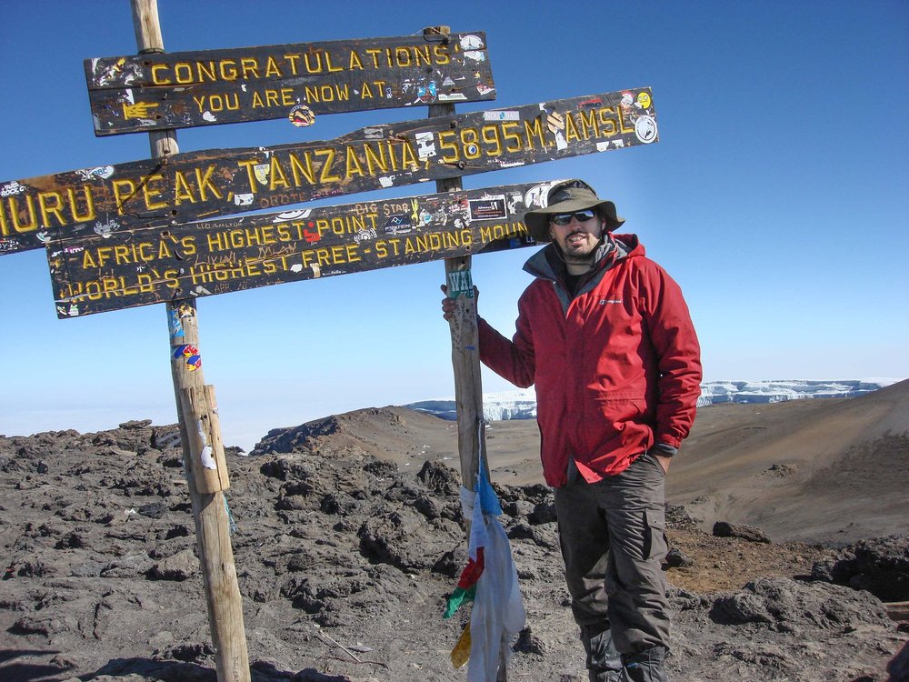 A man in a red coat and wide brimmed hat and sunglasses stand in a front of s sign that says he is at Africas highest point