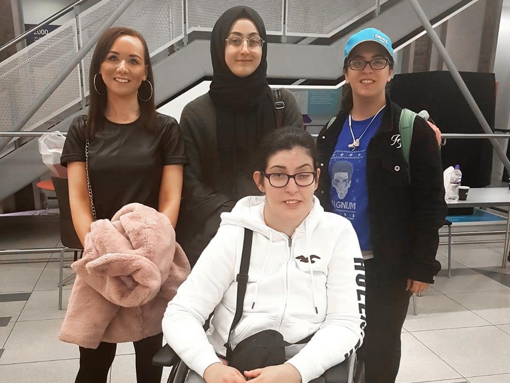 Three young women stand next to each other. In front of them is a women in a wheel chair