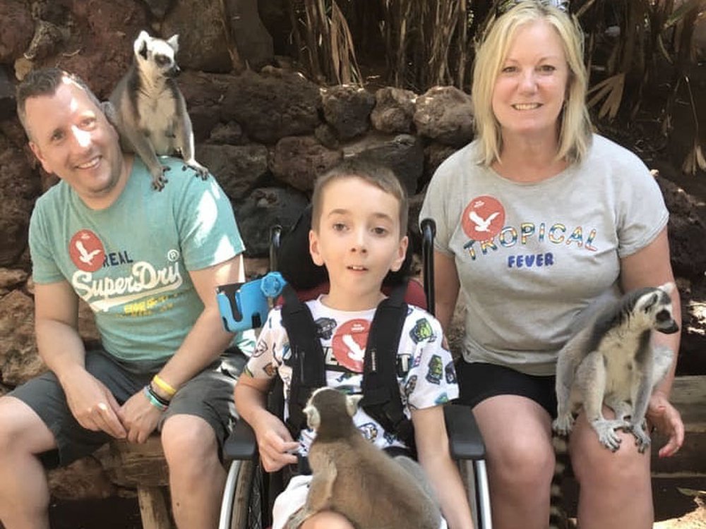 A man little boy and women smile into the camrea. Each have a lemur sat on them and the little boy is in a wheelchair