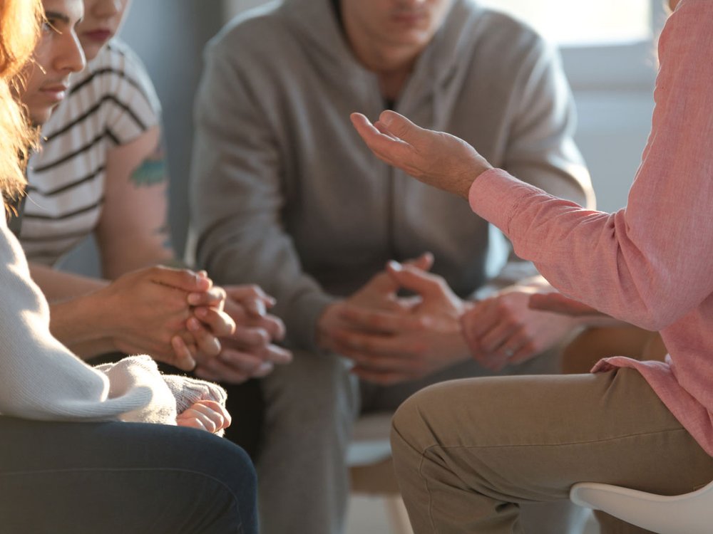 A group of adults sat all looking at one adult who is expressively holding their arms out